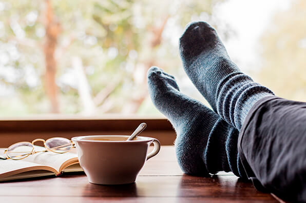 Wooden coffee table with cup of tea, book and spectacles and person's feet with blue socks on resting on table
